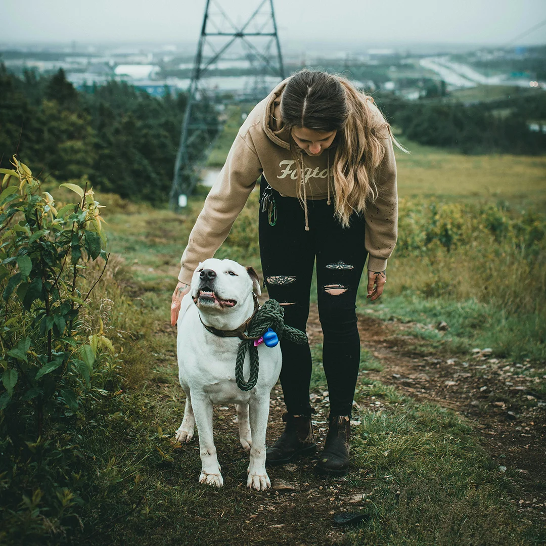 Woman in a tan hoodie petting a white dog with a rope leash while standing on a grassy trail beneath power lines.